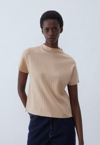 Beige ribkový pletený top s vysokým výstřihem a krátkými rukávy, s volnějším střihem a měkkou texturou. Nosený s tmavými denimovými sukněmi.