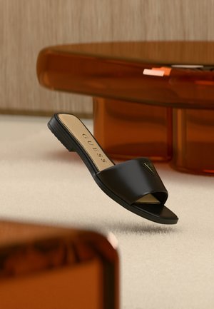 Black open-toe slide sandal with Guess logo on strap, resting on light carpet near curved amber-colored furniture.