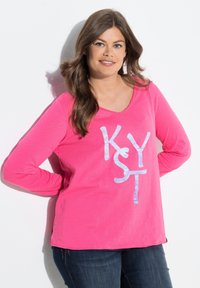 Rosa långärmad skjorta med V-ringning och en blanka vit grafik som säger "KYST." Lätt lös passform och rundad nederkant, tillsammans med mörka jeans.