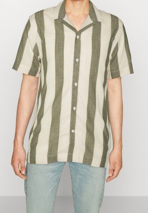 Hombre con una camisa de manga corta abotonada con amplias rayas verticales en verde oliva y beige, combinada con jeans azul claro.