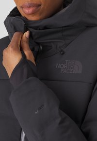 Giacca nera isolata con colletto alto, chiusura con zip e logo North Face in finitura opaca. Tessuto liscio e resistente all'acqua.