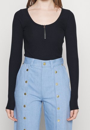 Femme portant un haut noir côtelé à manches longues avec une fermeture éclair sur le devant et un pantalon bleu clair taille haute avec des boutons dorés.