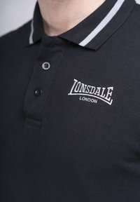 Camisa polo negra con una tela texturizada, que presenta un tapeta de dos botones y un cuello a rayas blancas. Logo bordado "LONSDALE LONDON" en el pecho.