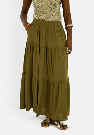 Mujer con falda larga de varias capas en color verde oliva, blusa de estampado paisley, sandalias doradas, pulsera dorada y anillo, con las manos en los bolsillos de la falda.