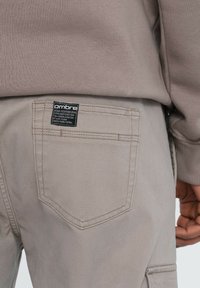Hintere Tasche von hellbeigen Hosen mit schwarzem Ombré-Label, getragen mit taupefarbenem Sweatshirtärmel und Hand sichtbar an der Seite.