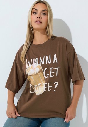 Mujer con cabello rubio largo lleva una camiseta marrón de gran tamaño con una ilustración de una taza de café y el texto "Wanna get coffee?" en letras blancas.