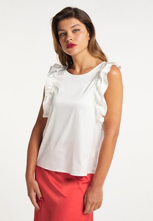 Jonge vrouw met bruin haar draagt een wit mouwloos topje met ruches en een rode rok, staand tegen een effen witte achtergrond.