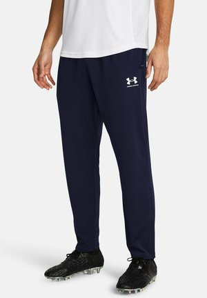 Parte inferior del cuerpo de una persona que lleva pantalones deportivos azul marino de Under Armour, camiseta blanca y tachones negros, de pie sobre un fondo blanco.