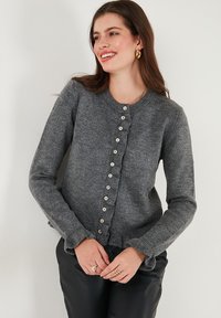 Cardigan grigio a maglia con scollatura tonda, bottoni frontali e orlo arricciato. Presenta maniche lunghe e una consistenza morbida.