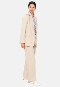 Completo beige su misura con giacca monopetto e rever, abbinato a pantaloni a gamba larga coordinati. Le sneakers bianche completano il look.
