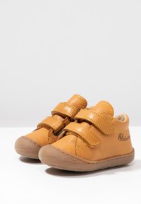 Kožené hnědé boty s dvěma suchými zipy, kulatou špičkou, gumovou podrážkou a viditelnými stehy. Logo potištěné na boku.