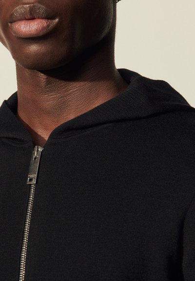 Sweat à capuche zippé noir avec un tissu texturé, détail de fermeture éclair argentée et cordon de serrage à la capuche. Gros plan sur le col et l'encolure.