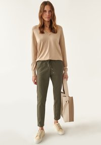 Suéter de punto beige con escote en v combinado con pantalones verdes oliva de cordón con detalles de botones, usados con zapatillas beige y una bolsa de mano.