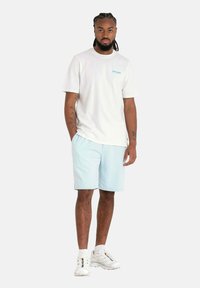 Witte t-shirt met blauw logo, lichtblauwe shorts, witte sneakers en schouderlang, donker haar, staand tegen een effen achtergrond.