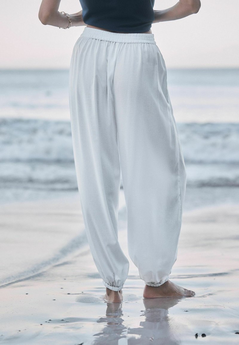 Person steht barfuß auf nassem Sand am Strand, trägt lockere weiße Hose und ein schwarzes ärmelloses Oberteil, mit Händen an den Hüften.