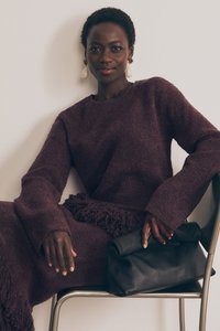 Lilla tekstuuriga sviiterikleit fringi detailiga ja pikkade varrukatega, koos musta nahast portfelliga. Modell istub metalltoolil.