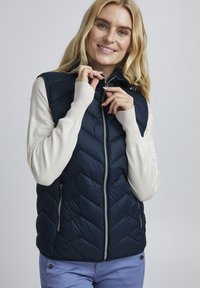 Gilet matelassé bleu marine avec motif en chevrons, fermeture éclair et poches latérales, porté sur un haut à manches longues beige clair.