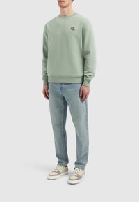 Lichtgroene sweatshirt van zacht materiaal, met een ronde hals en subtiele borduursels aan de voorkant. Gecombineerd met lichtblauwe jeans en sneakers.