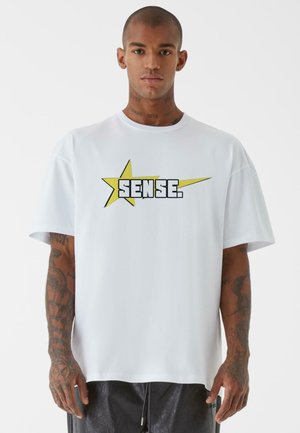 Bílá oversized tričko vyrobené z bavlny, s černou a žlutou hvězdou a nápisem "SENSE." potištěným přes hruď.