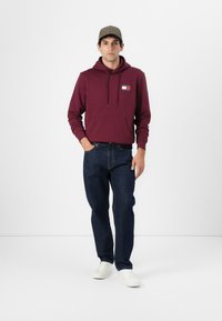 Bordeaux hættejakke med frontlomme og logo, parret med mørkeblå jeans og hvide sneakers. Modellen bærer en ternet kasket. Neutral baggrund.