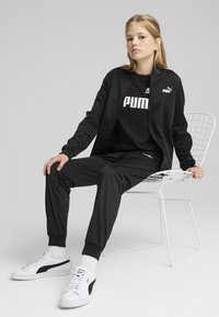 Ensemble de sport noir avec détails de logo, comprenant une veste zippée et un pantalon fuselé. Baskets blanches avec accents verts. Design minimaliste.