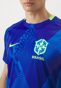 Homme portant un maillot de football du Brésil bleu avec le logo Nike jaune néon et l'emblème CBF sur la poitrine, sur un fond blanc uni.