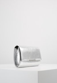 Silvermetallisk rektangulär clutchväska med en framficka och en liten metallplåt med varumärke, placerad på en vit pelare mot en enkel bakgrund.