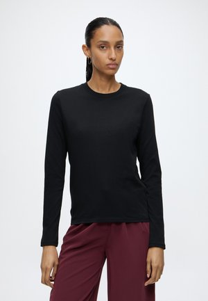 Femme debout devant un fond uni, portant un haut noir à manches longues et un pantalon bordeaux, tournée vers l’avant avec une expression neutre.