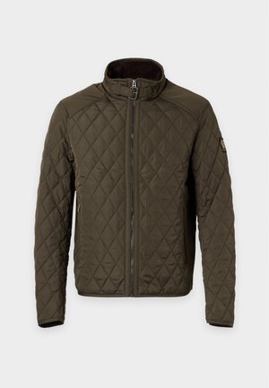 Homme portant une veste matelassée olive, un haut zippé bordeaux, un pantalon beige à jambes larges et des chaussures crème, debout sur un fond blanc uni.