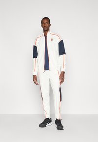 Αθλητική φόρμα σε λευκό χρώμα με ναυτικά σημάδια και πορτοκαλί ρέλι, περιλαμβάνει μπουφάν με φερμουάρ και αντίστοιχες παντελόνες, συνδυασμένη με μαύρα αθλητικά παπούτσια.