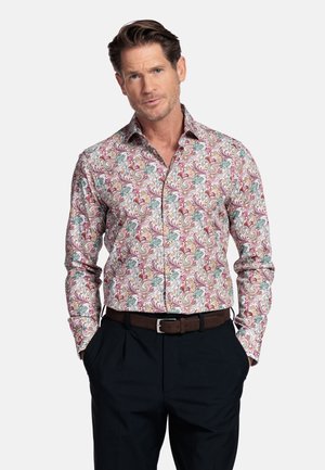 Man met een lange mouwen en een shirt met bloemenpatroon, ingestopt in een donker broek met een bruine riem, handen in de zakken, neutrale achtergrond.