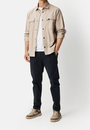 Homme portant une chemise beige boutonnée sur un t-shirt blanc, pantalon foncé et baskets beiges, debout avec une main dans la poche.