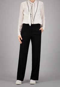 Blouse blanche transparente avec des bordures noires et un détail à nouer, associée à un pantalon noir taille haute. Des baskets blanches à enfiler complètent la tenue.