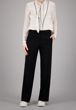 Witte doorschijnende blouse met zwarte bies en strikdetail, gecombineerd met hoge zwarte broek. Witte instappers ronden de outfit af.
