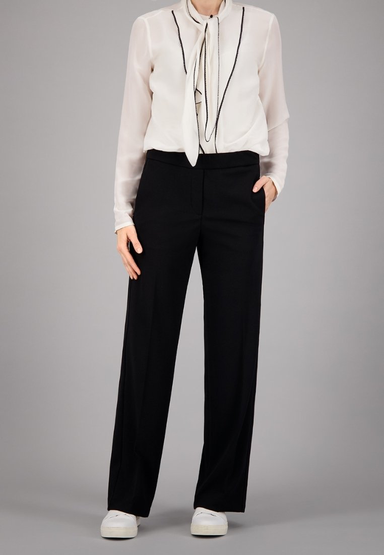 Blouse blanche transparente avec des bordures noires et un détail à nouer, associée à un pantalon noir taille haute. Des baskets blanches à enfiler complètent la tenue.