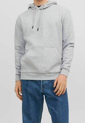 Mand iført lys grå hættetrøje med frontlomme og blå jeans, stående med afslappet kropsholdning mod en neutral baggrund.