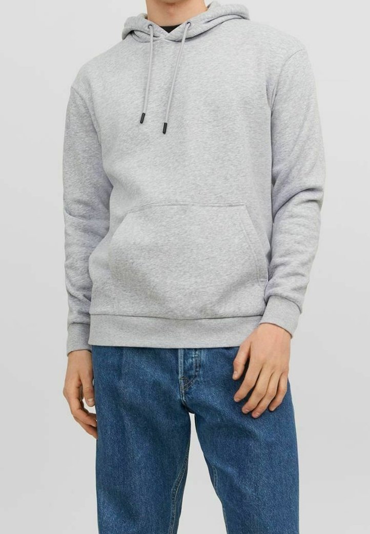 Homme portant un sweat à capuche gris clair avec une poche avant et un jean bleu, debout dans une posture décontractée contre un fond neutre.