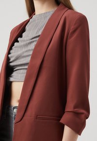 Rust-colored blazer with a tailored fit, notch lapels, and rolled cuffs. Features a smooth texture and a discreet front pocket.