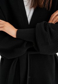 Mustat ribbikardiganit, joissa on kietaisuosa, leveät hihat ja solmittava vyö. Materiaali on tekstuuriltaan, mikä tuo syvyyttä vaatteeseen.