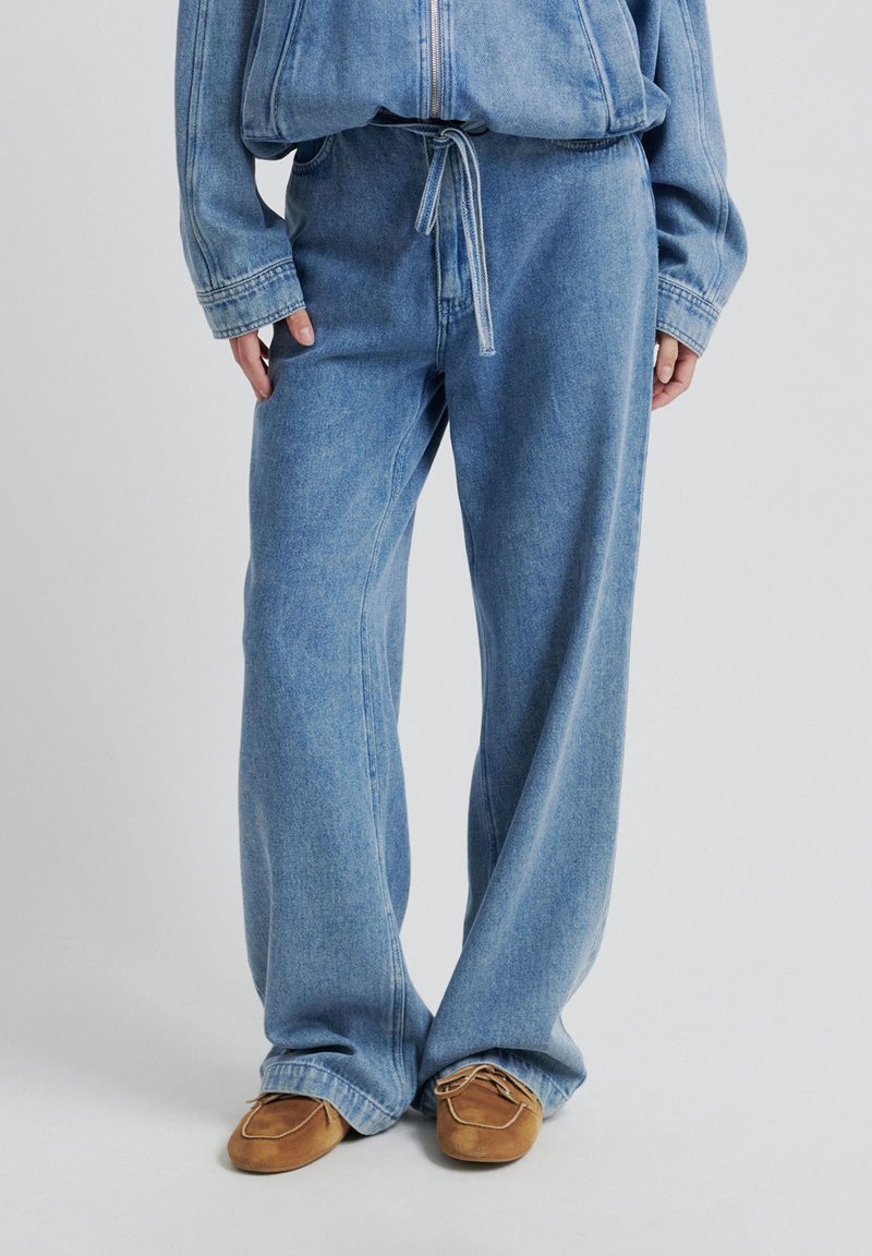 Široké kalhoty z světle modrého denimu se stahovací šňůrkou v pase, s viditelným prošitím a jemnou texturou. Na nohou jsou hnědé semišové boty.