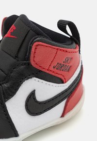 Svart, rød og hvit sneaker laget av lær og tekstilmaterialer. Har en høyt design, synlig søm og en løkkestropp. Merket "SKY JORDAN."