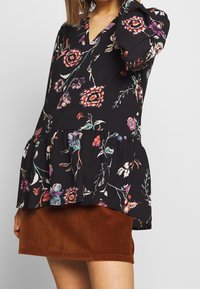 Schwarzes Blumen-Peplum-Oberteil mit Puffärmeln, V-Ausschnitt und mehrfarbigen Blumenmustern. Kombiniert mit einem braunen Cordrock.