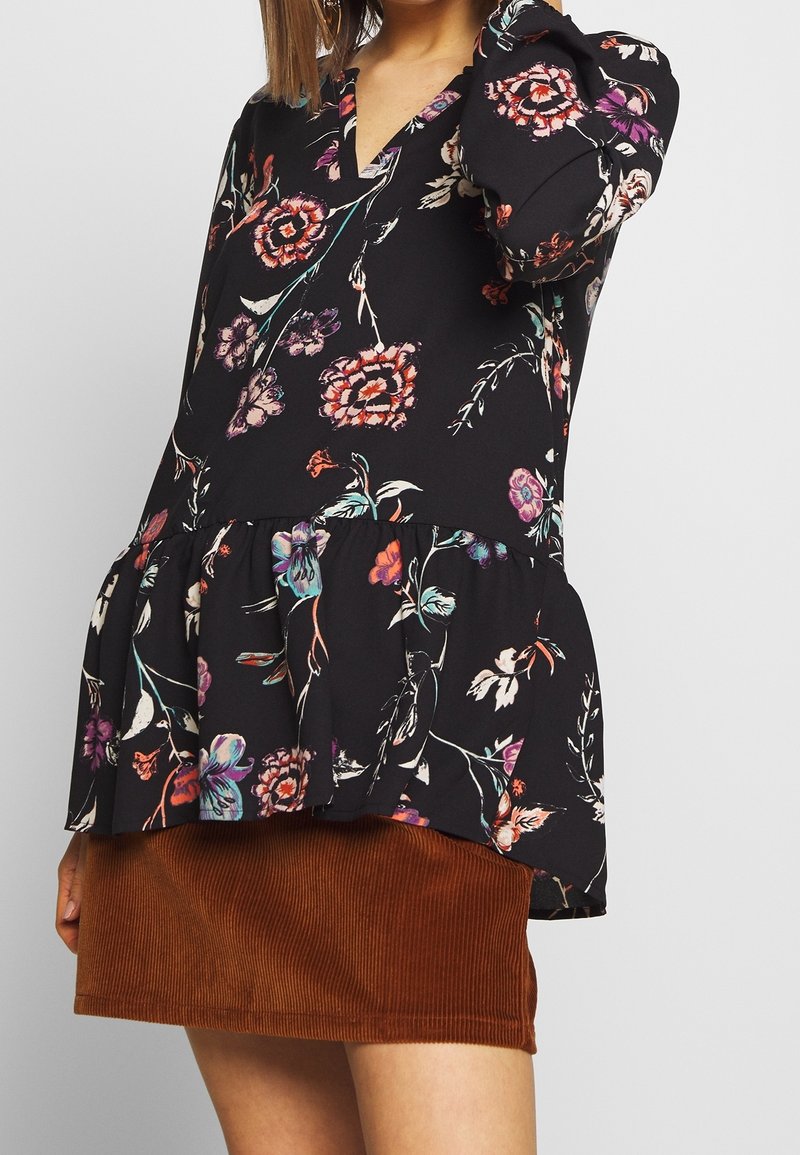Schwarzes Blumen-Peplum-Oberteil mit Puffärmeln, V-Ausschnitt und mehrfarbigen Blumenmustern. Kombiniert mit einem braunen Cordrock.
