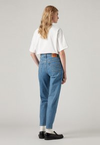 Kvinna med långt blont hår, iklädd en vit T-shirt, blå högmidjade jeans, vita strumpor och svarta loafers, stående på sidan mot en neutral bakgrund.