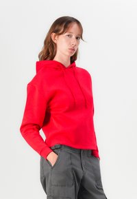 Sarkans kapucēts džemperis, kas izgatavots no mīkstas auduma, ar samazinātu piedziņu, priekšējo kabatu un auklu. Parasts ar pelēkiem cargo biksēm.