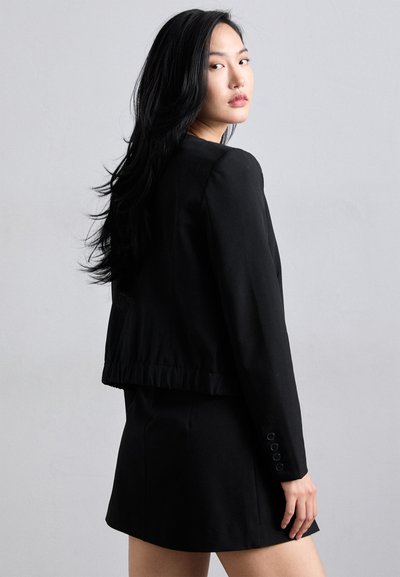 Black cropped blazer with a smooth texture, featuring a gathered hem and buttoned cuffs. Worn over a matching black mini skirt.