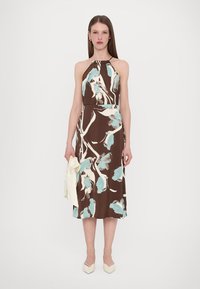 Robe fleurie marron avec des motifs bleu clair et blanc, design à encolure halter, longueur midi et texture douce. Le mannequin tient une pochette blanche.
