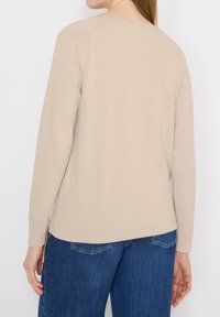 Femme portant un pull beige à manches longues et un jean bleu en denim, vue de dos sur un fond blanc uni.