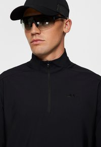 Giacca sportiva nera con zip e colletto alto, dotata di un piccolo logo sul petto. Accompagnata da un cappellino nero e occhiali da sole scuri.
