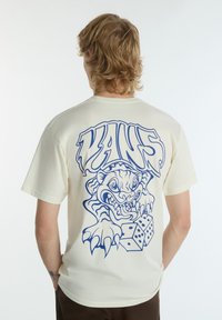 Cremefarvet T-shirt med en stor blå grafik af et monster, der klorer på terningerne på ryggen. Kortærmet og afslappet pasform.